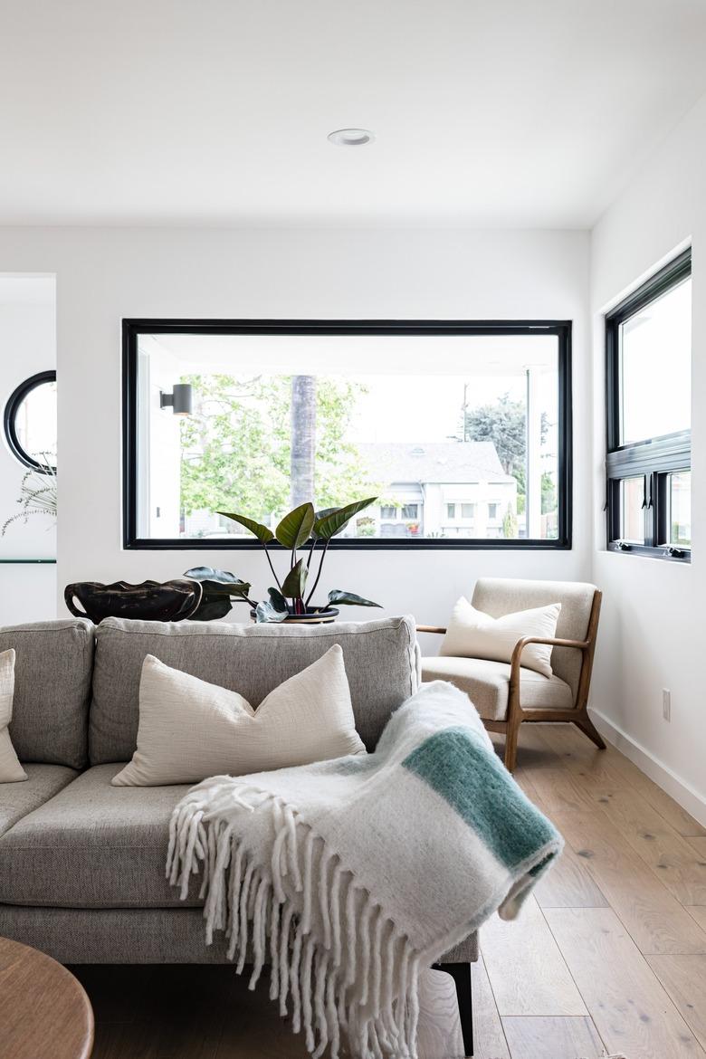 Grey couch with white fringed blanket in modern living room with large black-framed windows and hardwood floor