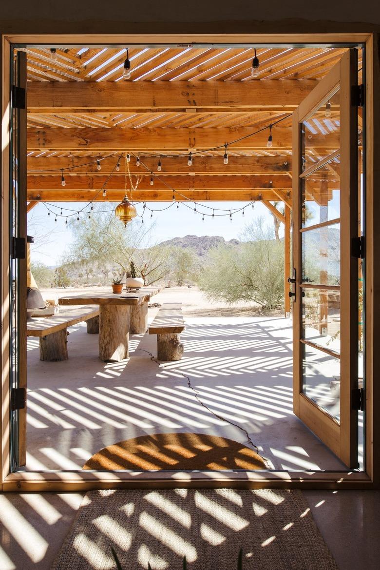 Open French doors, leading to a wood pergola with string lights. A wood picnic table with plants and an ochre pendant light is above.