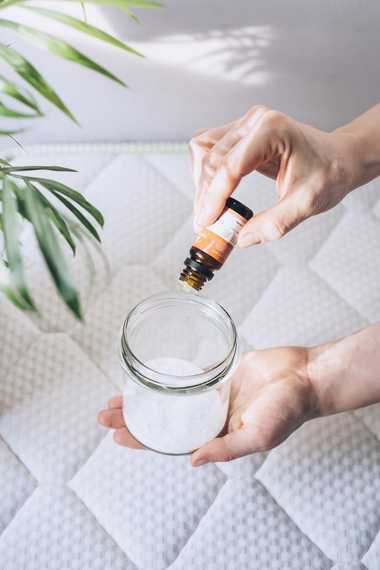 Hand pouring baking soda into small glass container over white mattress with white headboard next to plant