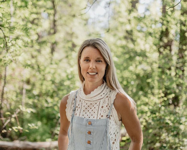 Anne Helen Petersen in a white shirt and overalls in front of green trees.