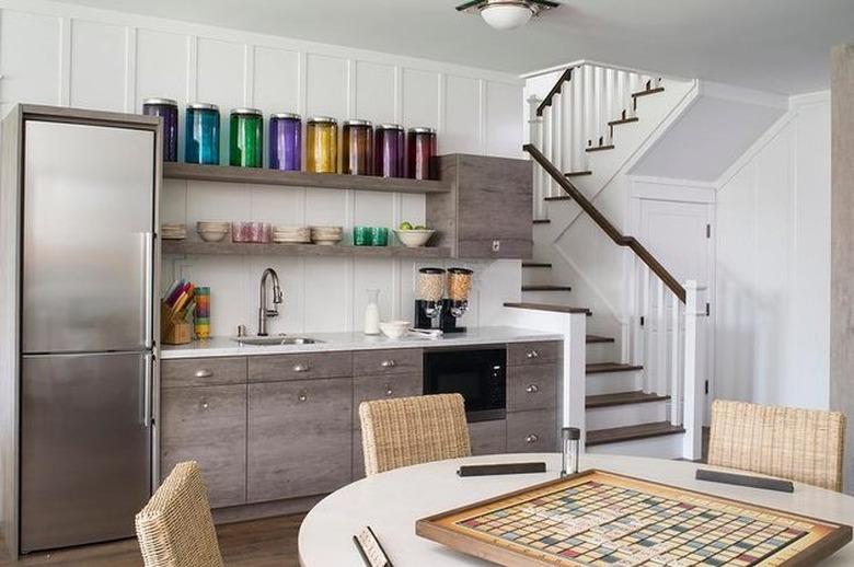 basement ideas with Stainless steel fridge and gray wood cabinets with open shelves, sink, round card table, stairway.