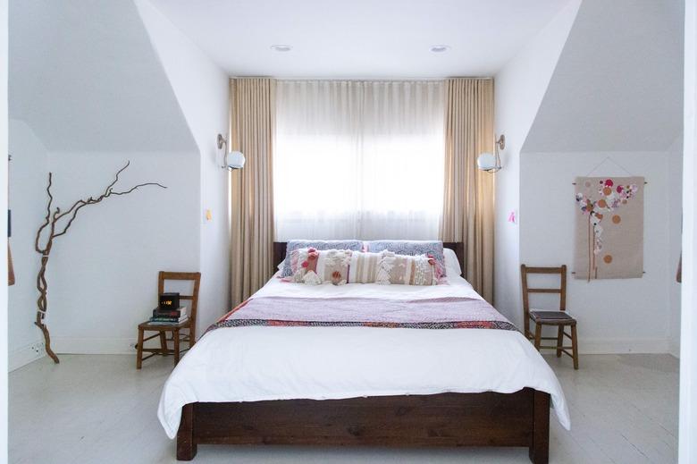 A bedroom with neutral bedding and layered window treatments over the window