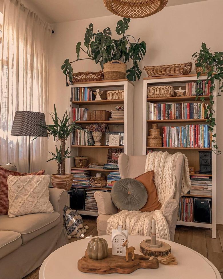 A beige living room with two white bookshelves behind a beige couch and chair draped with a white blanket and a couple pillows.