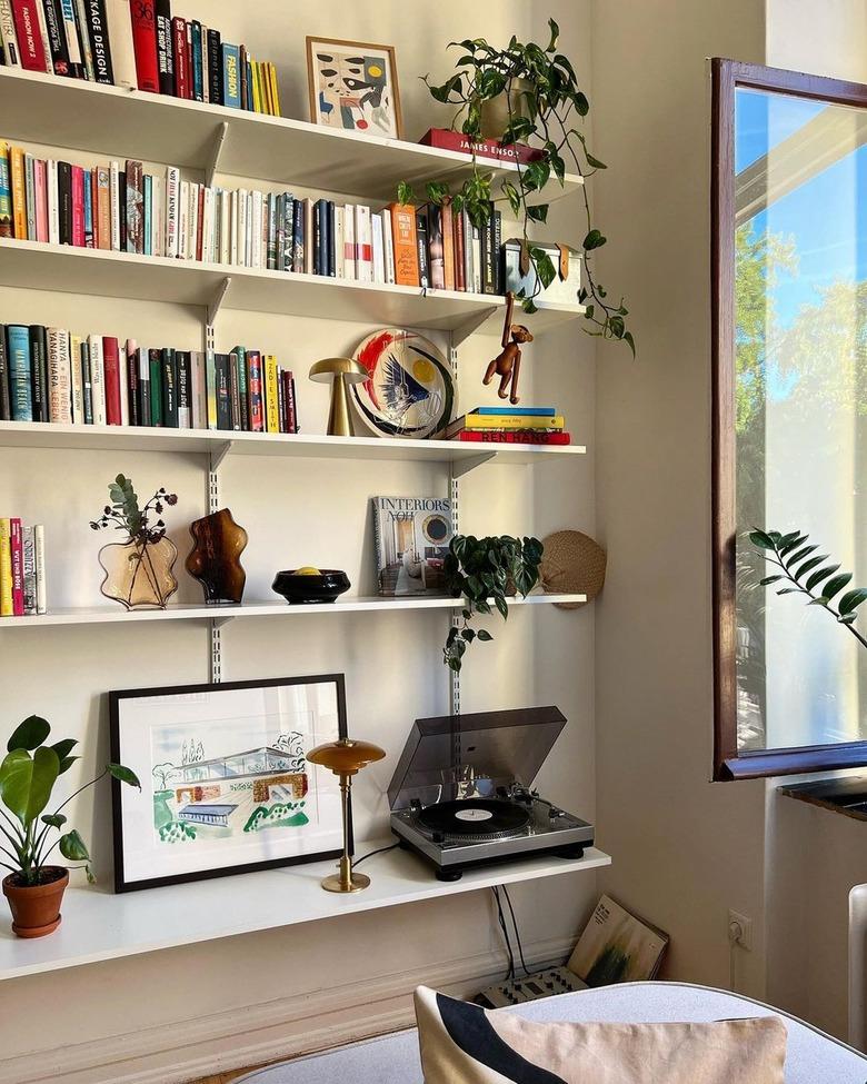 A minimalist bookshelf made with single shelves hammered into the wall next to an open window.