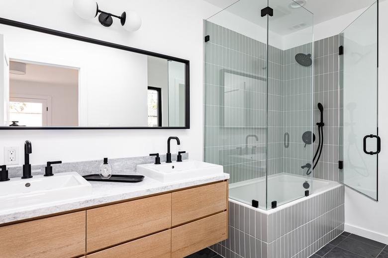 Modern bathroom with glass-walled shower over white-tiled bathtub, black faucets and shower head, white sinks, and black-framed mirror