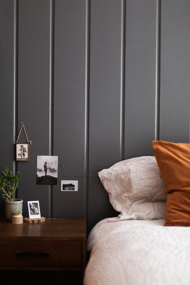 Bedroom with a gray wainscoting wall and small photos. Wood nightstand with plants and a bed with brown and neutral pillows.