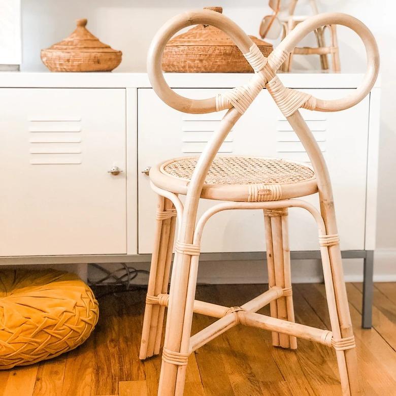 A woven chair with a bow shape at the top in front of a white dresser on a wooden floor.