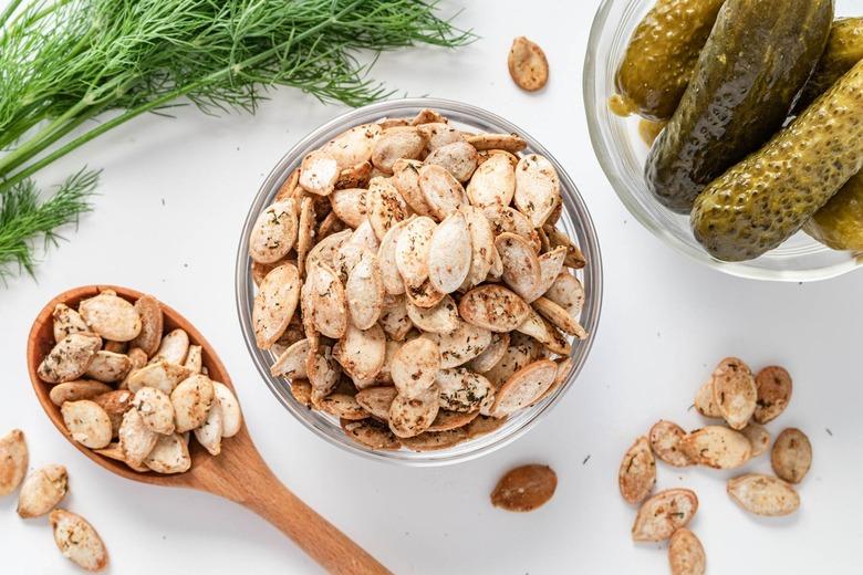 A glass bowl of dill pickle seasoned pumpkin seeds surrounded by a glass bowl of dill pickles and fresh dill.