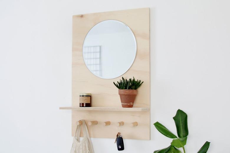 a shelving unit made from a sheet of plywood with a shelf, several thick wooden pegs, and a mirror at eye level