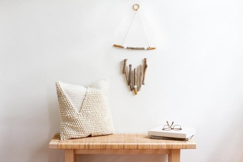 diy rustic decor wall hanging, made of several vertical pieces of driftwood suspended from a horizontal one, hangs above a wooden bench with a rattan top