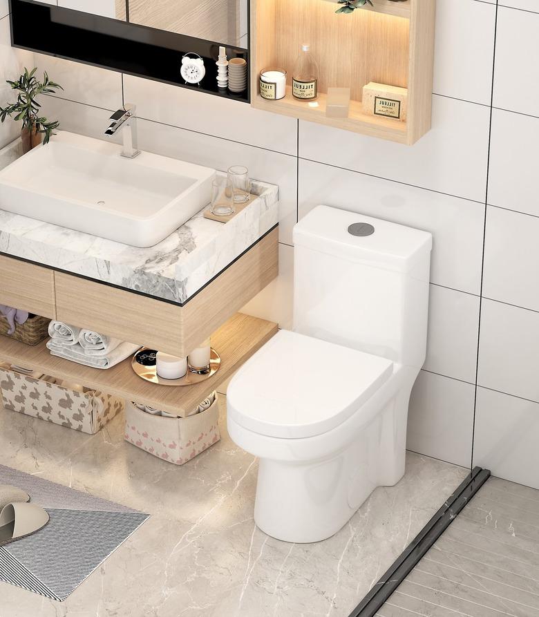 A white dual-flush toilet in a bathroom with floating shelves and large white tile on the walls