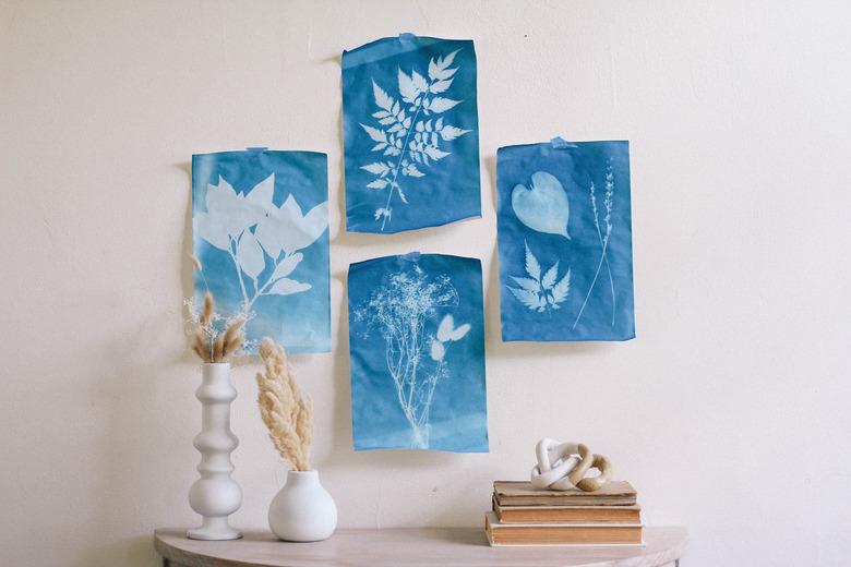 Four blue sun prints hung on wall with tape above table with white vases and books