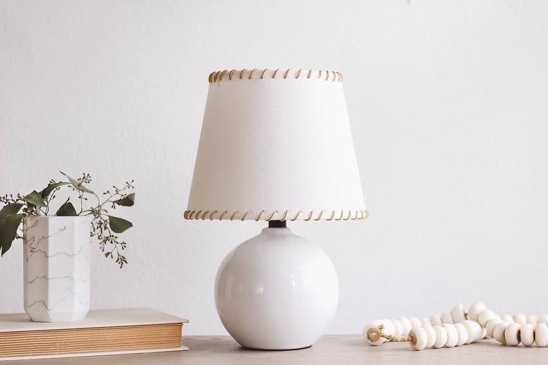 SKOTTORP lampshade with leather whipstitching, on a table with a book, and a vase of flowers