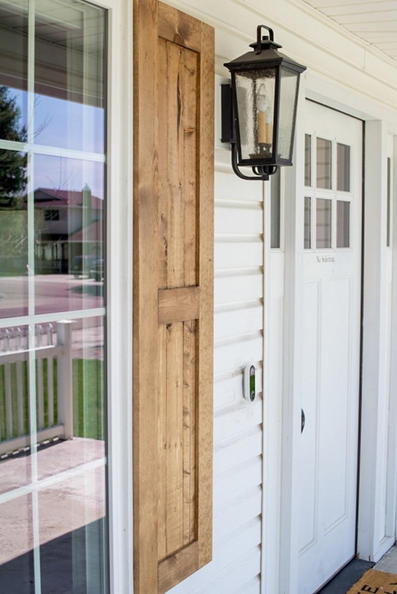 Flat panel exterior shutter style in natural wood finish on white exterior next to lantern sconce