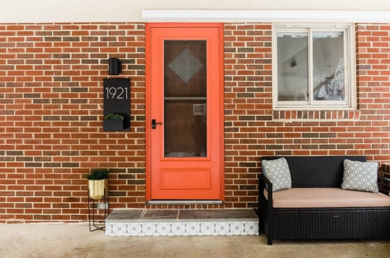 Brick house with an orange door, black wicker bench, gold plant stand, and black sconce light