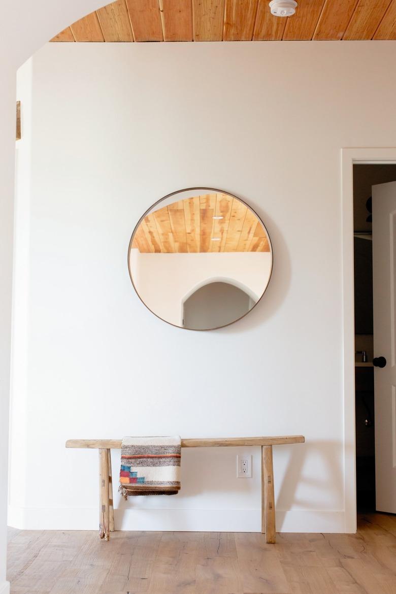 Hallway Furniture Ideas with A circular mirror on a white wall. Underneath, there's a low wooden bench with a throw blanket hanging on it.