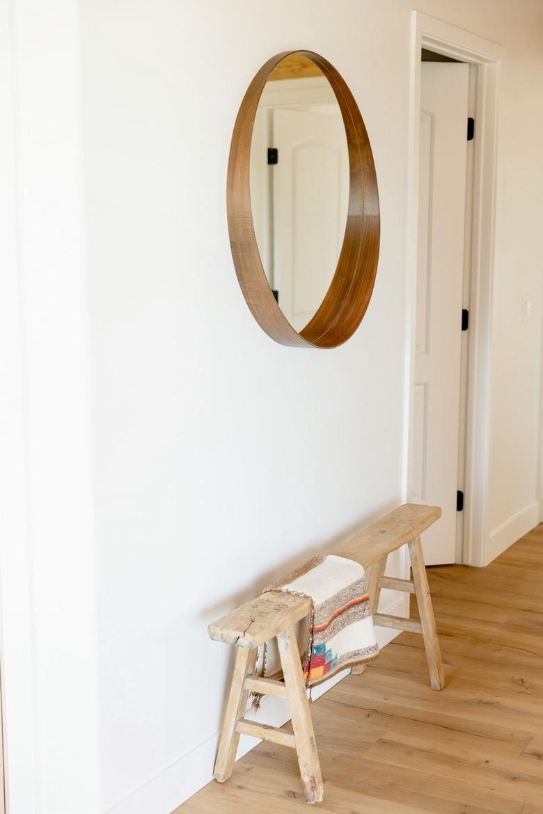 A circular mirror on a white wall. Below it, a low wooden bench with a decorative throw.