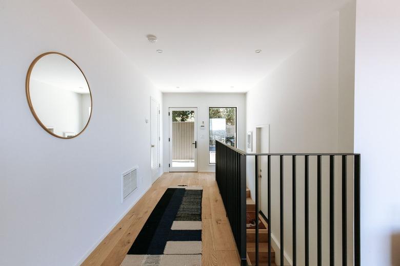 A hallway with light wood floor with an ornate red rug, a round wall mirror, casement windows and black banister staircase