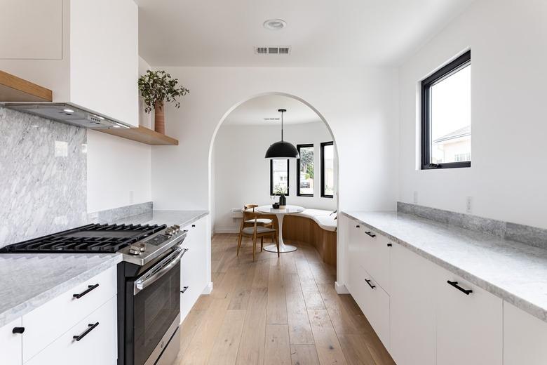 View of dining room with curved bench, small round table, chairs, pendant lamp, and tall windows through archway from white-walled kitchen space