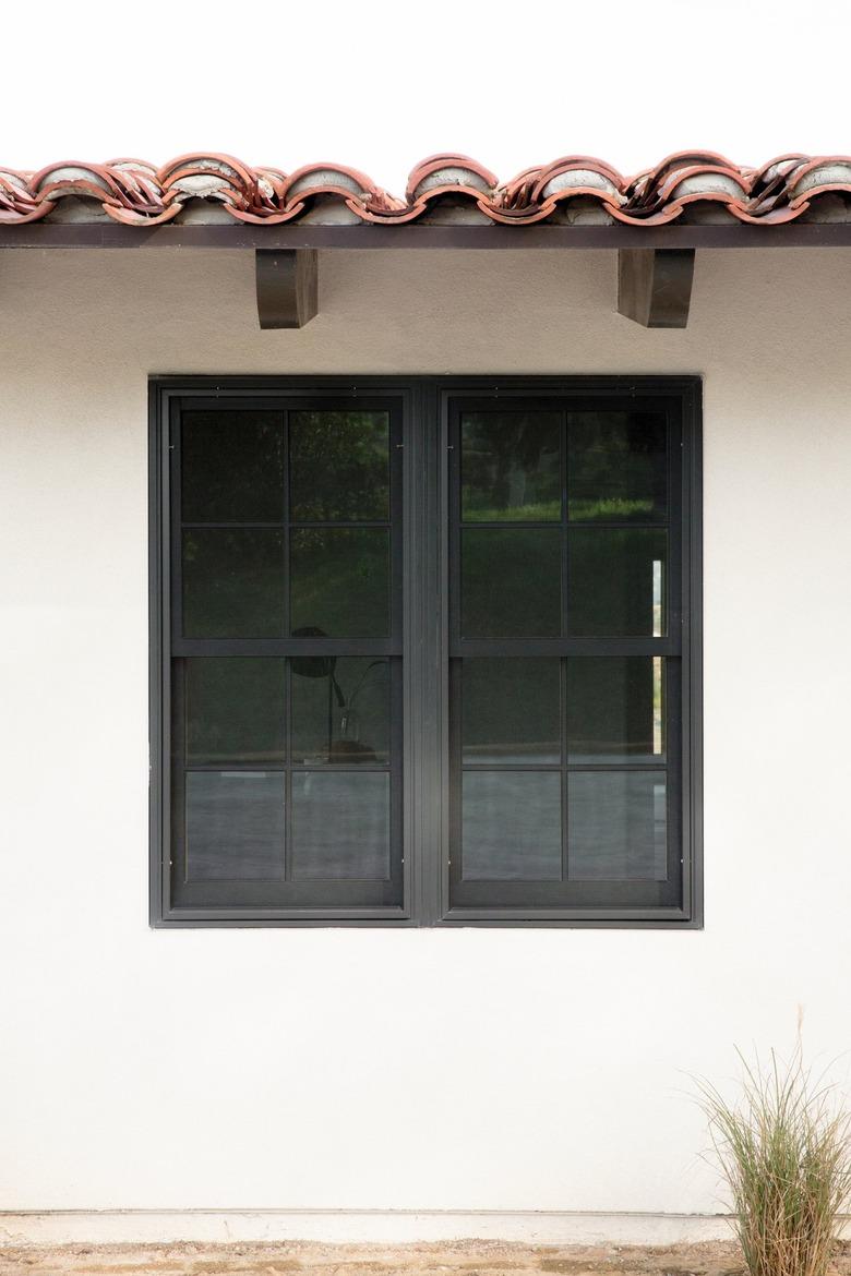 Side of a Spanish-style home with white walls and a clay tile roof with dark wood support beams. There is a rectangular window in the middle of the wall. On the ground, a small Mexican feather shrub.