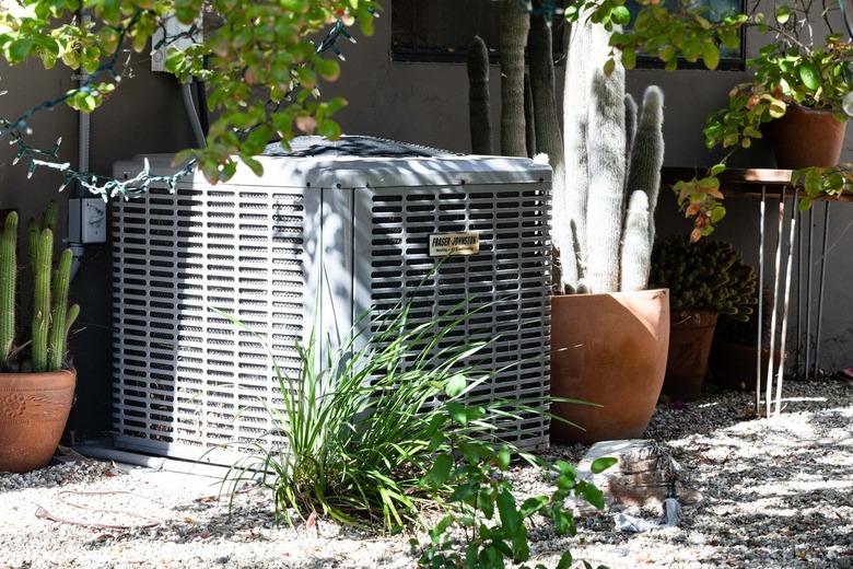 A gray cube A/C unit in a shady spot surrounded by outdoor plants.