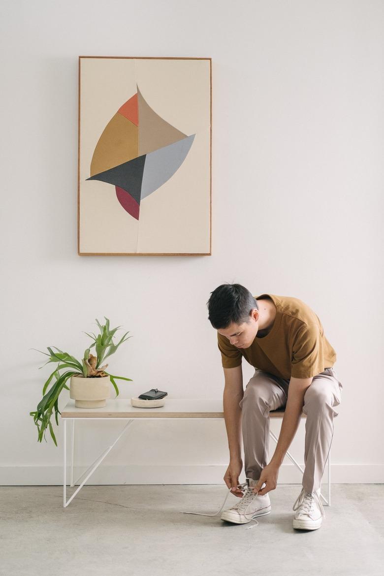 Man sitting on bench with potted plant tying shoes against white background with canvas art piece