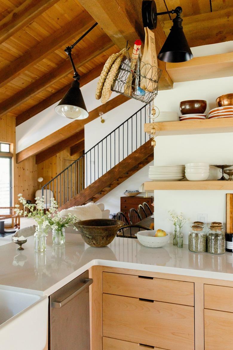 Kitchen with wood shelves, white counter, wood cabinets. Vases of flowers and black lamp sconces. A wood staircase in the distance.