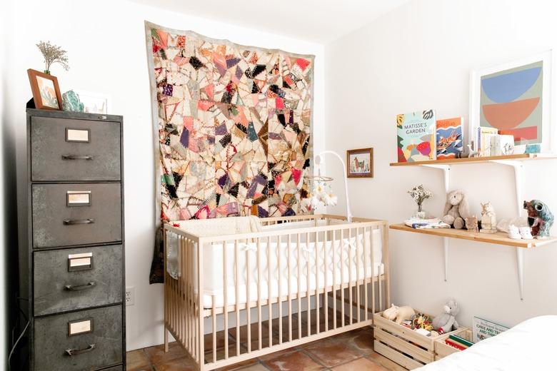 Nursery with a wood crib and baby mobile. Colorful quilt hangs beside. Wood shelves and crates with toys and books, and a metal cabinet.