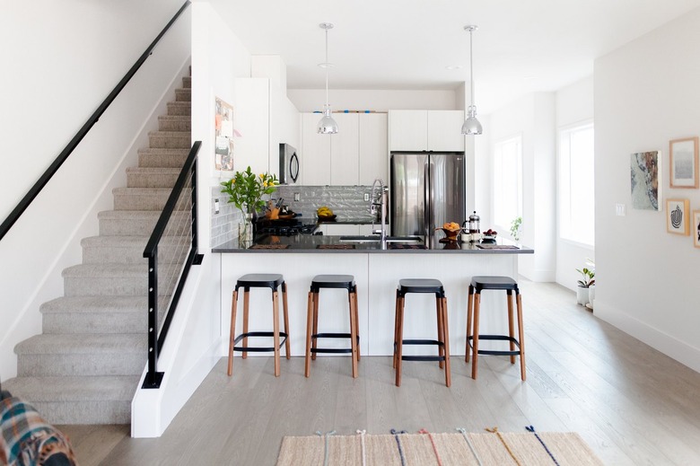 Wood stools at a gray counter kitchen island, with gray pendant lights. A kitchen with a gray backsplash. A gray carpeted staircase.