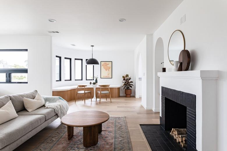 Modern living room with couch, rug, fireplace, and coffee table leading to dining room with pendant lamp, curved bench, and table set