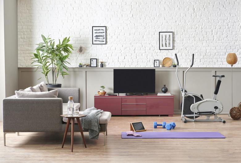 Man is doing sport and looking cell phone training program. Decorative living room, television unit, grey sofa purple mat and blue dumbbell brick wall home interior concept.