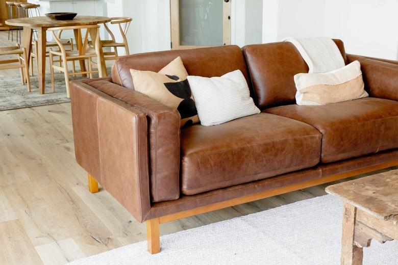 A brown leather sofa with three pillows and a white throw blanket. The sofa is on a white rug, and a wood coffee table is visible. Behind the sofa, there is a light wood table with four chairs and a dark brown bowl in the center of the table.