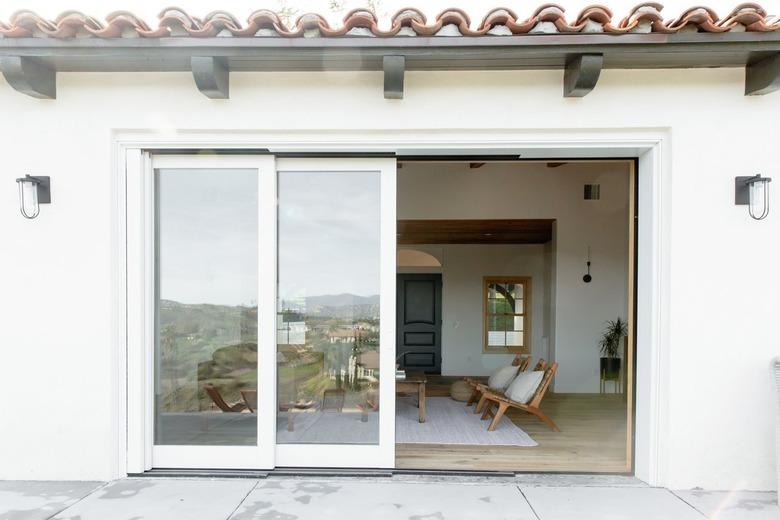 Glass sliding doors with white frame leading to a living room where two teak chairs with accent pillows and a rustic coffee table are visible.