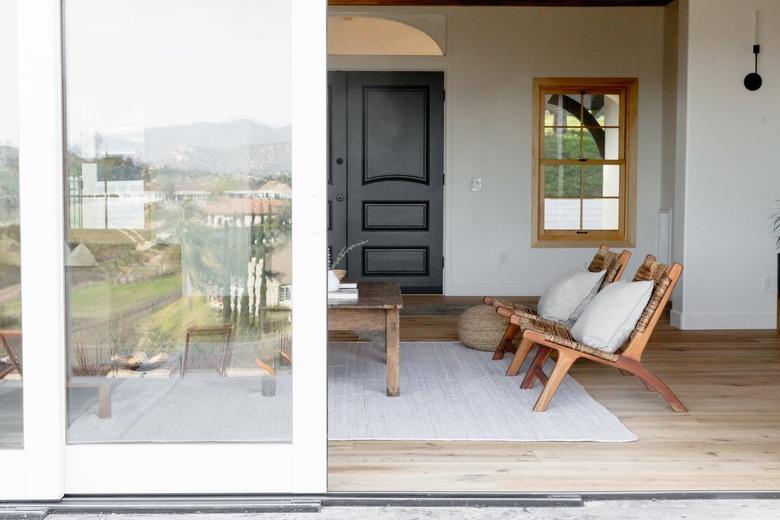 Glass sliding doors with white frame leading to a living room where two teak chairs with accent pillows and a rustic coffee table are visible.