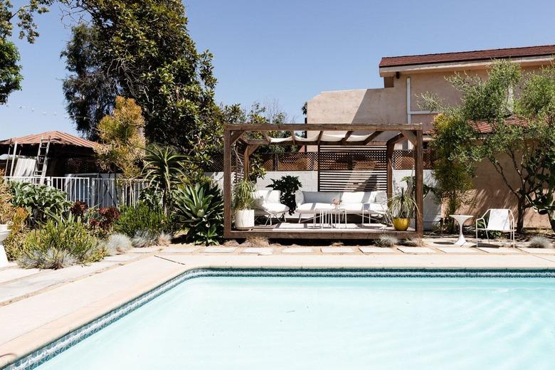 Swimming pool with a wood pergola with white patio furniture and tropical plants.