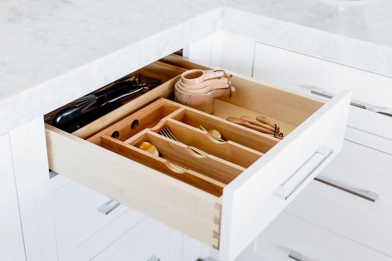 An open drawer with a wooden cutlery organizer. Cutlery, measuring cups and spoons, and other utensils visible inside. Marble countertop above the drawer.