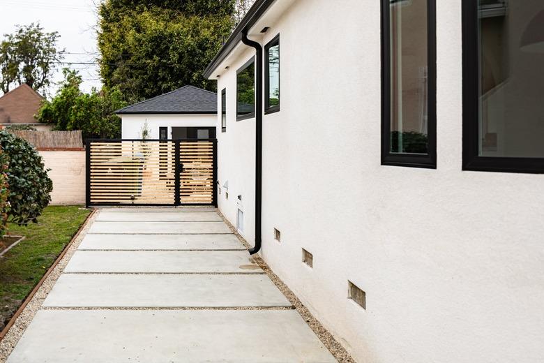 Outdoor walkway alongside of white-walled house with large concrete tiles leading to black fence with grass and bushes