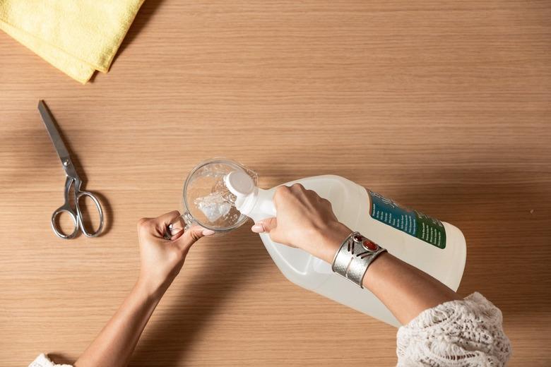 Hand pouring liquid from plastic jug into measuring cup next to scissors and yellow dryer sheets against wood countertop