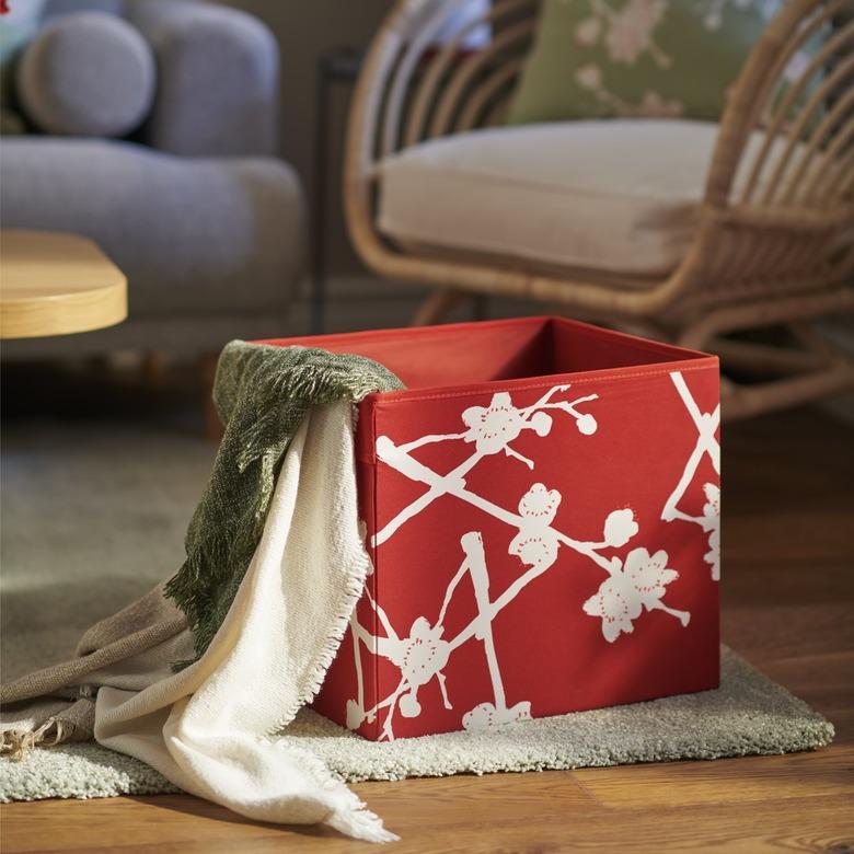 A red box with a white floral pattern full of blankets on a rug in a living room.