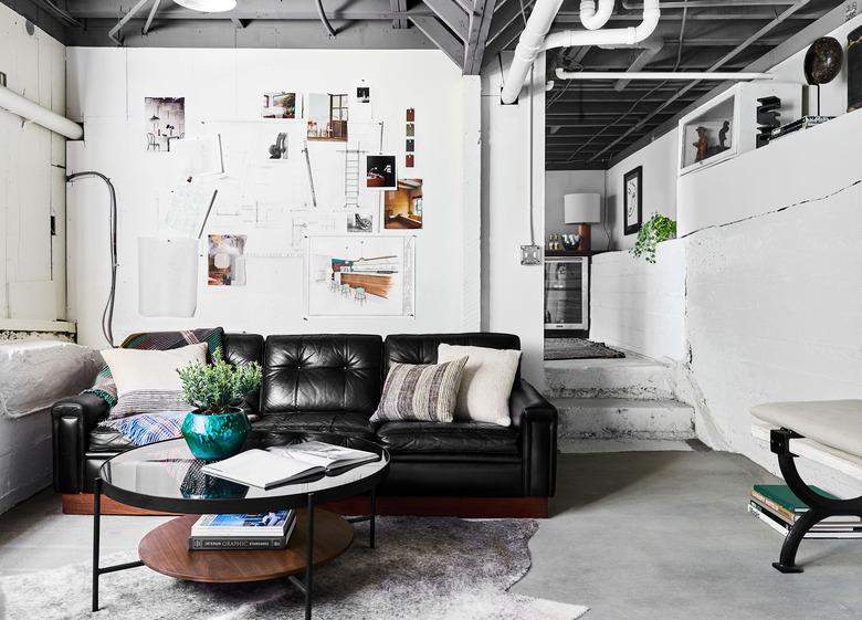 White walls, concrete floors, black leather couch, exposed ceiling, round coffee table, and cowhide rug in industrial basement