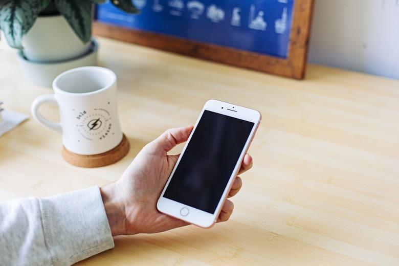 Hand holding an iPhone over a wooden surface next to a white coffee mug on a brown coaster.