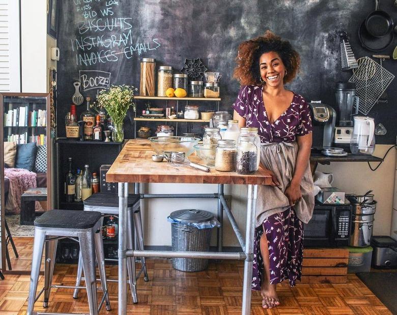 Jerrelle Guy in a purple floral dress sitting on a stool next to a wooden counter topped with mason jars of spices.