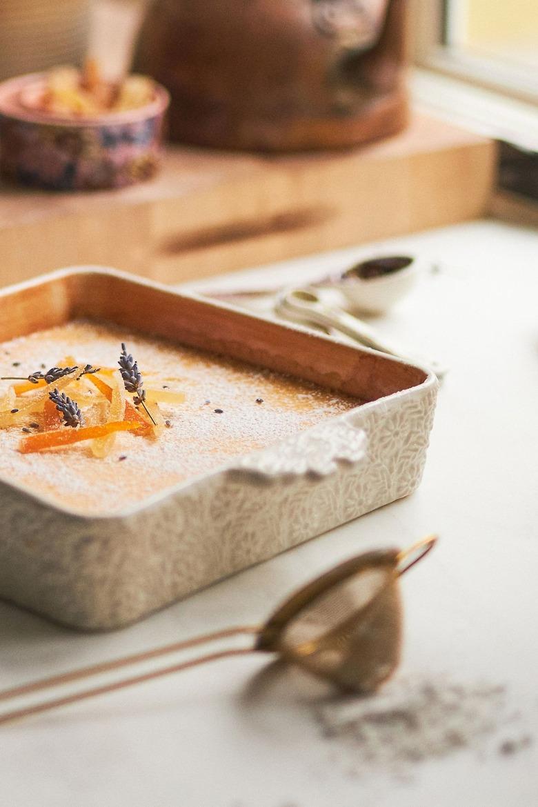 A cake with candied orange on top rests in a baking dish on a kitchen counter by a flour sifter.