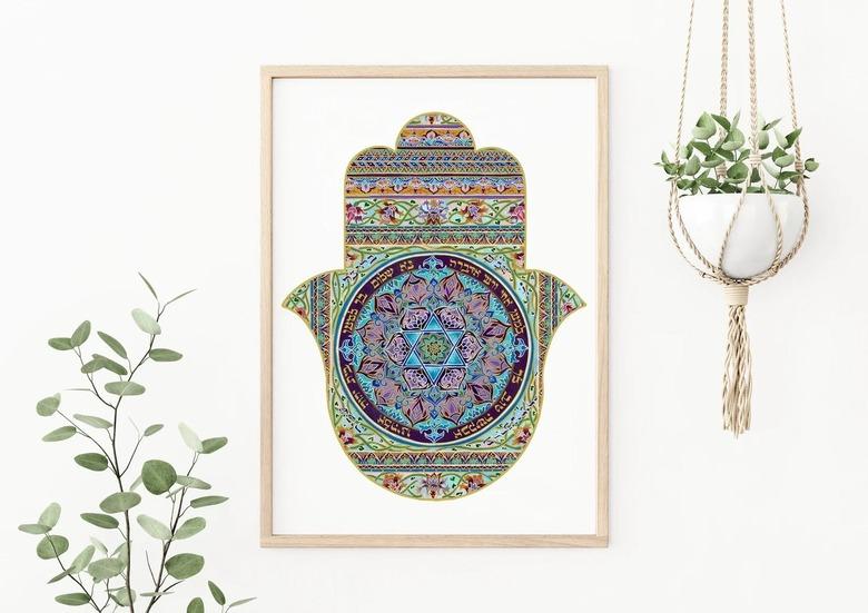 A patterned hamsa in a wooden frame hanging on a white wall next to a hanging plant.