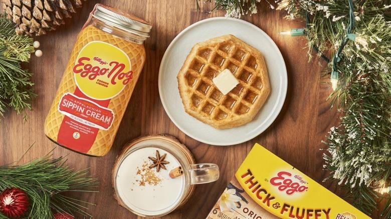 A holiday flat lay: Garland, holiday lights and a pinecone surround a glass of eggnog, a box of Eggo Thick and Fluffy waffles, a waffle with syrup and butter on a white plate, and a jar of Eggo Nog Sippin' Cream rest on a wooden surface.