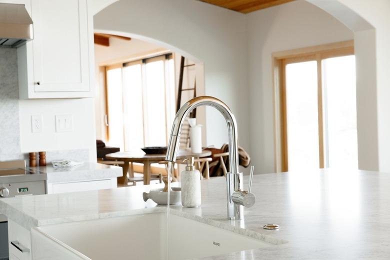 In a kitchen, a sink in a marble countertop, part of a kitchen island. The chrome faucet is running water. Next to the sink, a marble soap dispenser and a wooden scrub brush in a ceramic dish.