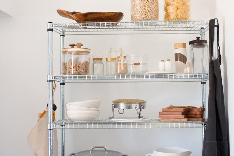 Metal shelf racks with clear containers of dry goods, white dish ware, wood cutting boards, and a gray cast iron pot.