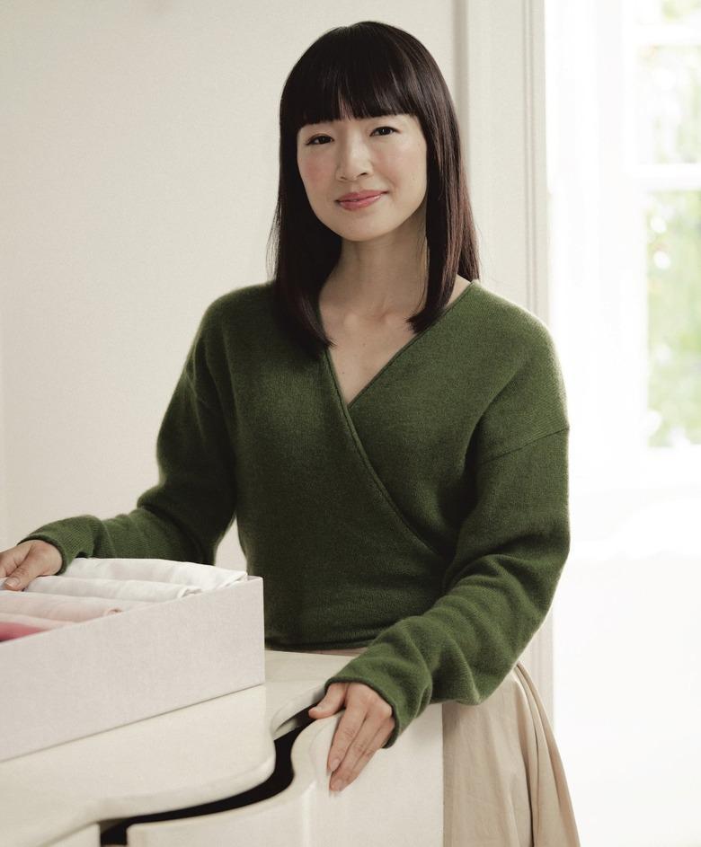 Marie Kondo in a green v-neck sweather and beige skirt in front of a white wall and window.