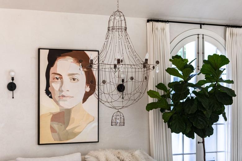 A white-walled living room with a tree plant, arched windows, a wire chandelier and painting