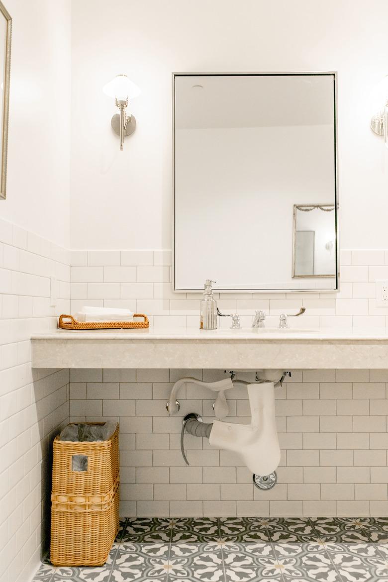 white bathroom with wall-hung sink, rectangular mirror,  subway tile wall, vinyl flooring with a floral pattern, rattan basket, light sconces